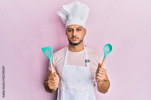 Young arab man wearing professional cook apron and hat holding spoon relaxed with serious expression on face. simple and natural looking at the camera.