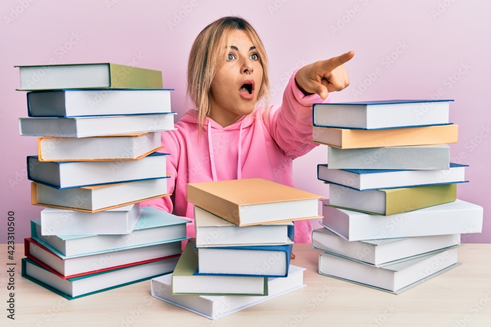 Young caucasian woman sitting on the table with books pointing with finger surprised ahead, open mouth amazed expression, something on the front Young caucasian woman sitting on the table with books pointing with finger surprised ahead, open mouth amazed expression, something on the front