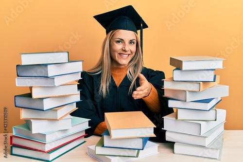 Young caucasian woman wearing graduation ceremony robe sitting on the table smiling friendly offering handshake as greeting and welcoming. successful business.