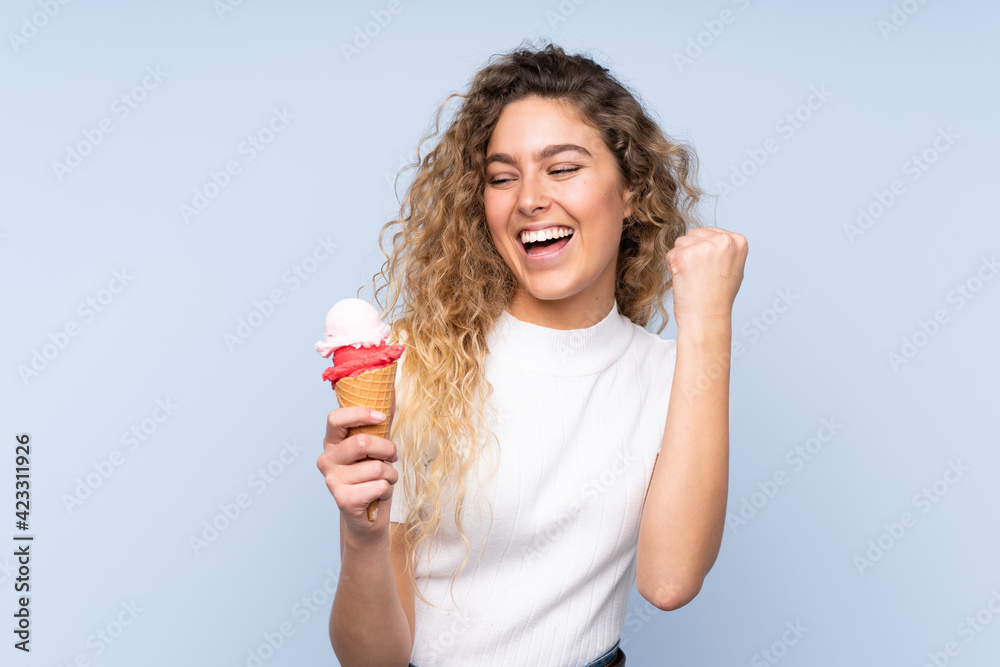Young blonde woman with curly hair holding a cornet ice cream isolated on blue background celebrating a victory Young blonde woman with curly hair holding a cornet ice cream isolated on blue background celebrating a victory