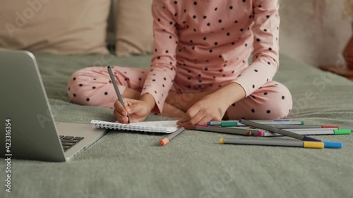 Cute little African American girl learning to draw with felttip pens by looking to laptop. Teenage girl in pajamas sits on bed in bedroom with beautiful light interior. Slow motion. Close up. photo
