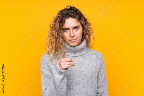 Young blonde woman with curly hair wearing a turtleneck sweater isolated on yellow background frustrated and pointing to the front © luismolinero