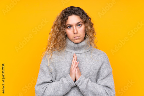 Young blonde woman with curly hair wearing a turtleneck sweater isolated on yellow background keeps palm together. Person asks for something © luismolinero