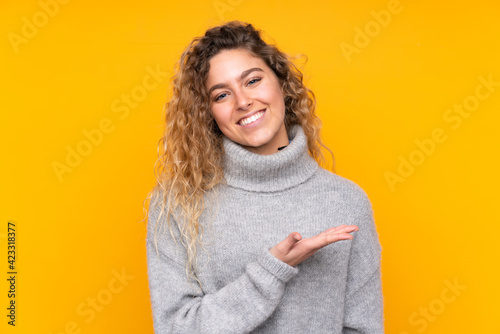 Young blonde woman with curly hair wearing a turtleneck sweater isolated on yellow background presenting an idea while looking smiling towards © luismolinero