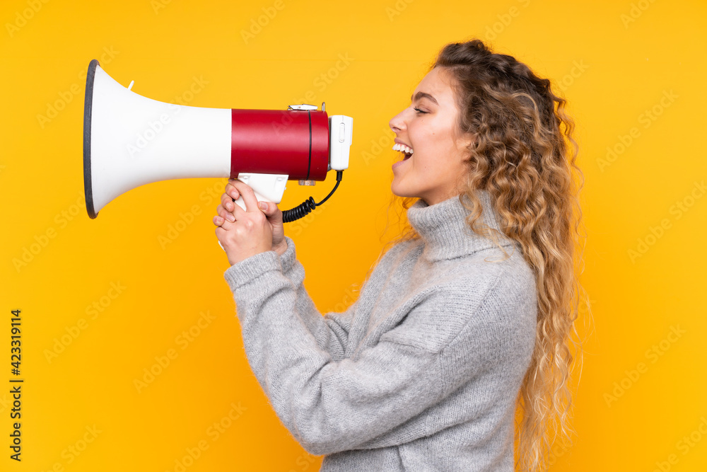 Young blonde woman with curly hair wearing a turtleneck sweater isolated on yellow background shouting through a megaphone Young blonde woman with curly hair wearing a turtleneck sweater isolated on yellow background shouting through a megaphone