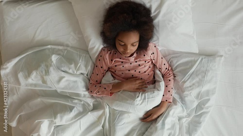 Top view of a little sleeping African American girl waking up stretching and yawning. Teenage girl in pajamas lies on white bed in bedroom. Slow motion. photo