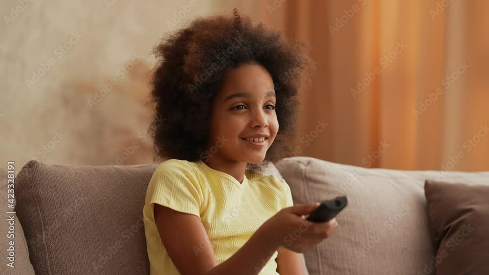 Portrait little African American girl watches TV and switches channels with interest with remote control. Teenage girl sits on gray sofa in hall. Slow motion. Close up.
