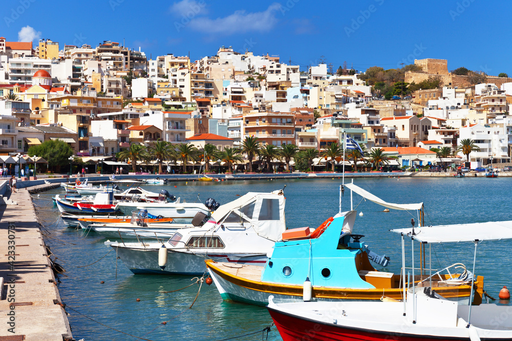Greece island Crete. Beautiful view of the town of Sitia on the hill and fishing boats on blue water near embankment on sunny day. Southern bright seascape. Summer seaside vacation and travel
