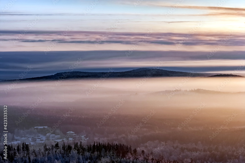 Fototapeta premium Winter landscape with forest, houses, mountains, haze, sky, clouds in the rays of the setting sun