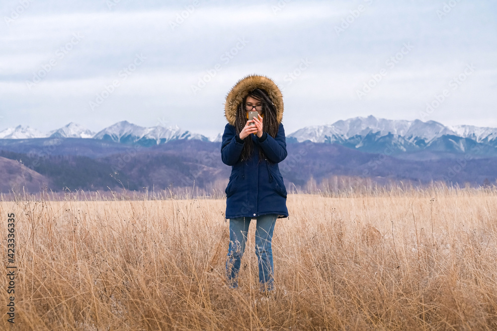 Human and nature. Caucasian girl with dreadlocks looks at her cell phone on snowy mountains background. Gadget addiction