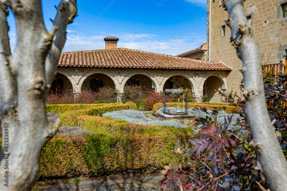 Obraz premium Monastery garden view with well with a rope, well maintained plants and typical church architecture. Meteora, Greece, sunny early spring day landscape