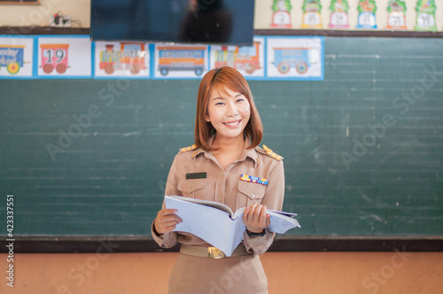 female thai teacher in government uniform standing in front of classroom reading an open note book smiling