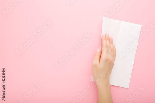 Young adult woman hand wiping light pink table background with dry white paper napkin. Closeup. Pastel color. General or regular cleanup. Empty place for text or logo. Top down view. Copy space.