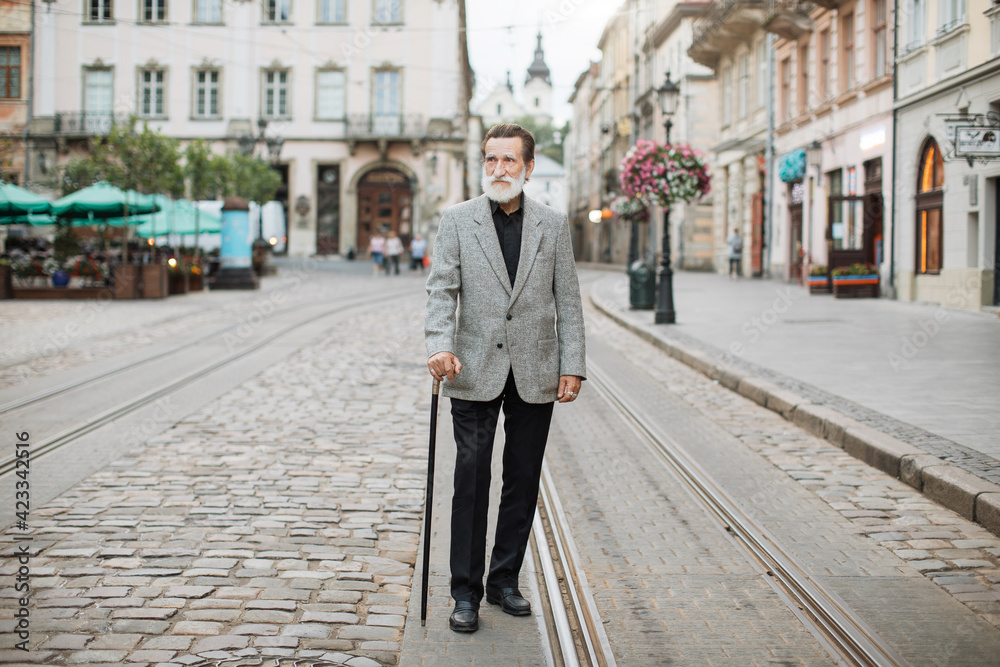 Senior man in stylish outfit walking on street with cane in hand. Caucasian pensioner with grey beard enjoying fresh air at old town.