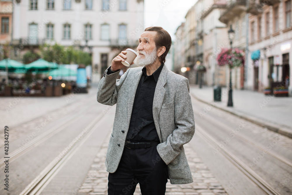 Confident stylish man with grey beard standing at city street with cup of fresh coffee in hand. Senior male walking outdoors and drinking hot beverage. Confident stylish man with grey beard standing at city street with cup of fresh coffee in hand. Senior male walking outdoors and drinking hot beverage.