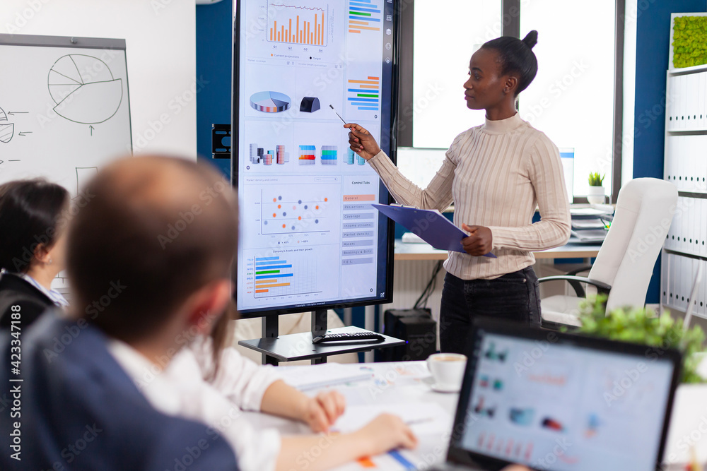 Black woman worker explaning company statistics in front of colleagues, briefing group of employees. Multiethnical businesspeople working in professional startup financial office during conference Black woman worker explaning company statistics in front of colleagues, briefing group of employees. Multiethnical businesspeople working in professional startup financial office during conference