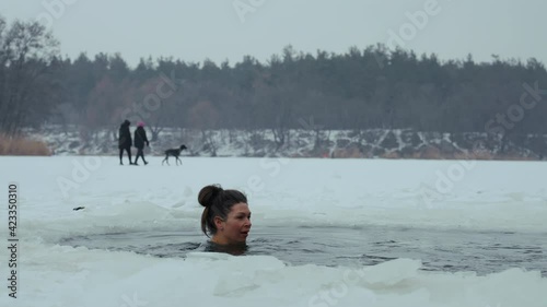 Woman with hair bun plunging in ice hole in frozen river, blurred people walking with dog on background. Young female enjoying winter traditions. Concept of extreme activities photo