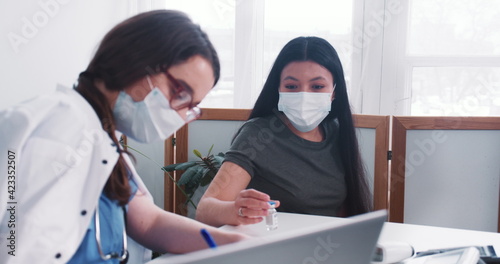 Young Caucasian doctor woman consults mixed race patient woman wearing mask  holding medication flask at medical exam.