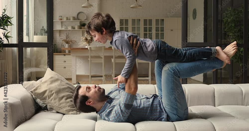 Strong cheerful young dad lying on comfy sofa in cozy living room lifts up on outstretched arms his little preschool son. Family practicing yoga, play having fun enjoy active weekend together at home