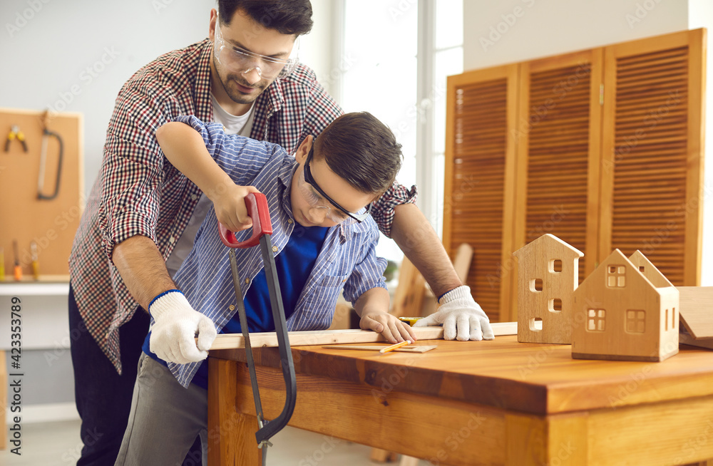 Father teaching son to use sharp handsaw sawing wood plank together at home carpentry workshop. Masculine duties, parent and child common hobby concept. Woodworking, family leisure activity Father teaching son to use sharp handsaw sawing wood plank together at home carpentry workshop. Masculine duties, parent and child common hobby concept. Woodworking, family leisure activity