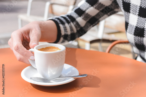 hand of a caucasian young woman holding a white cup of coffee on the top of an orange table at a cafe terrace