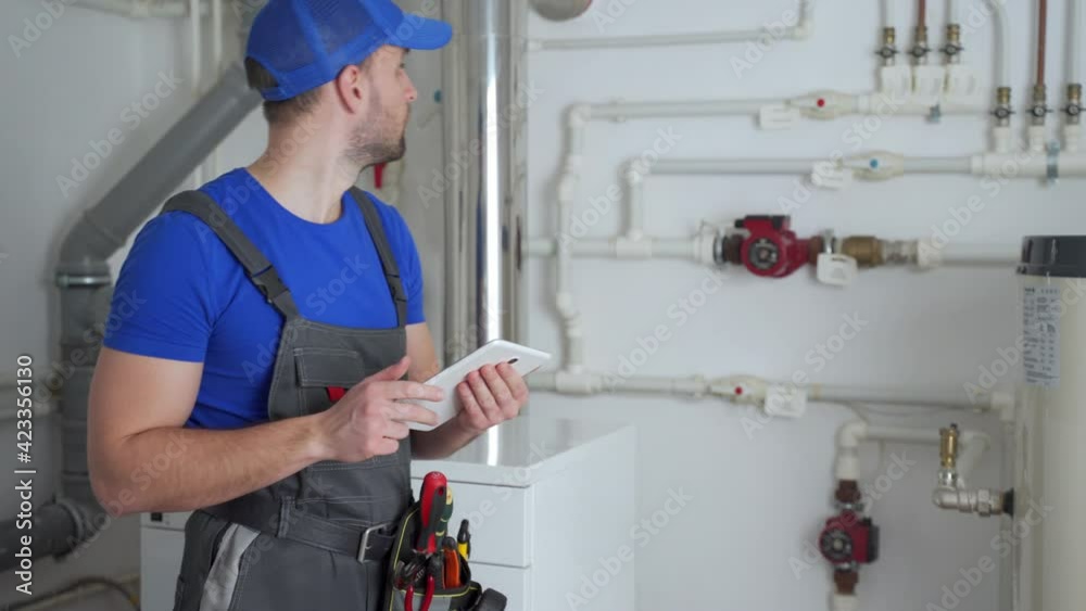 Young worker in a protective suit uses a tablet while checking the boiler equipment in the house.