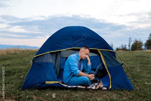 Man working on laptop in tent in nature. Young freelancer sitting in camp. Relaxing in mountains. Remote work, outdoor activity in summer. Happy male work on vacation.