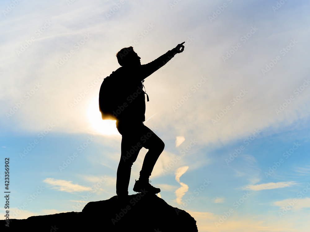Silhouette of a man with one arm raised and backpack. Pointing to the sky. Climber at the top of a mountain. Finished and celebrating the climb