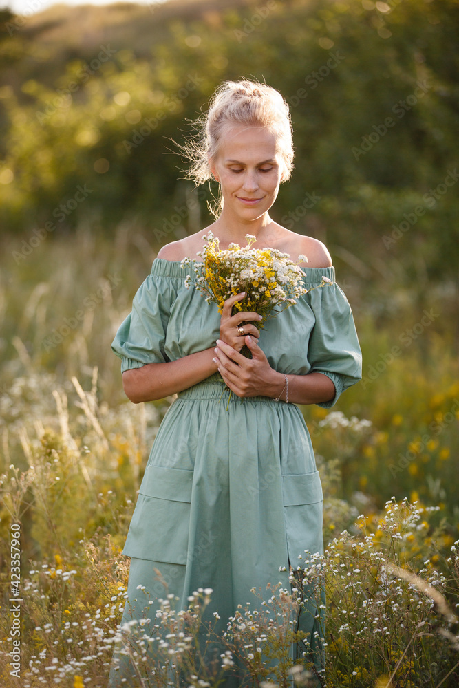 Summer mood. Romantic woman weared in long mint cotton dress walk in a meadow on background of flowering grass. Backlight. Spring or summer coming. Blonde-haired girl with bouquet of wild flowers Summer mood. Romantic woman weared in long mint cotton dress walk in a meadow on background of flowering grass. Backlight. Spring or summer coming. Blonde-haired girl with bouquet of wild flowers