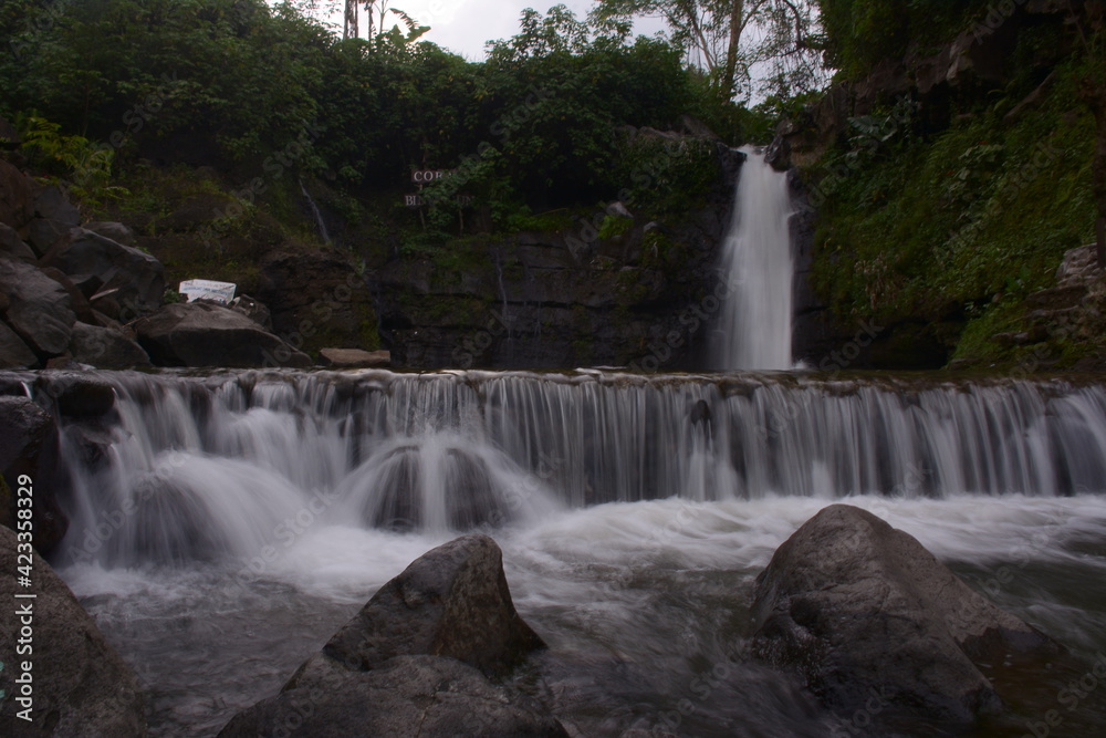 A stunning jungle waterfall flows into a cool mountain river with big rocks.