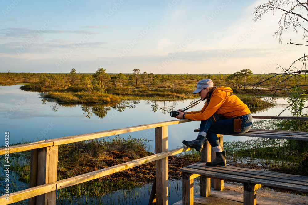 Woman photographer shoots the landscape of a forest lake among the raised bogs at sunset.