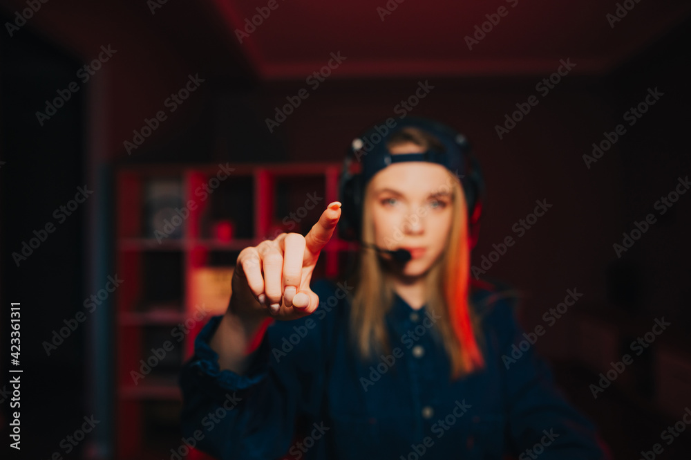 Obraz premium Close photo of a female gamer in a headset on her head showing a finger to the camera. The focus on the lady gamer's finger shows the screen.