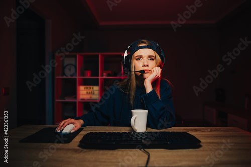 Tired female gamer in headset playing computer games at night at home, staring intently at camera.