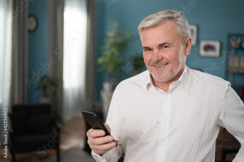 Close up face portrait of healthy cheerful mature gray-haired businessman with smart phone smiling to the camera while standing in the living room. Happy mature middle aged grandfather.