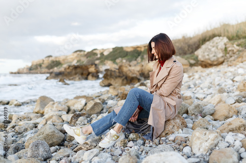 A beautiful young woman sits on a rocky shore and thinks about something. Evening portrait of a beautiful young woman in nature. Solitude, retreat, isoloation, seclusion concept