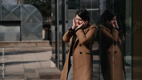 young woman dressed in a beige coat listening to music with her wireless headphones on outdoors