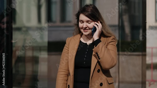 young woman dressed in a beige coat speaking on the phone explaining something outdoors