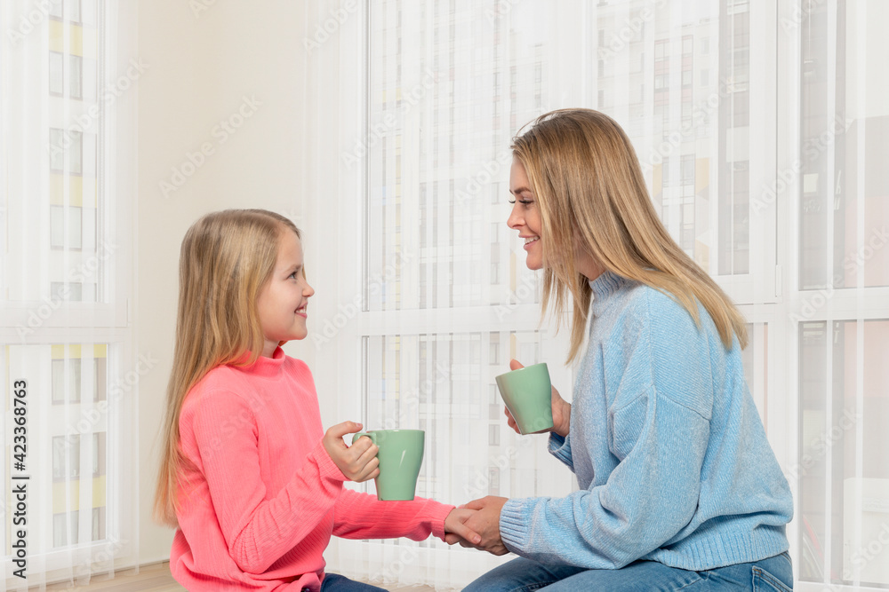 Fototapeta premium Young mother and daughter with cups of tea spends time together