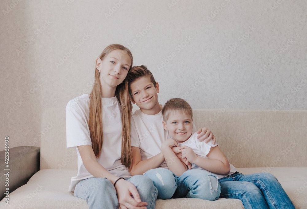 three children, a girl and two boys in white T-shirts and blue jeans, sit hugging each other on the home sofa and smile.