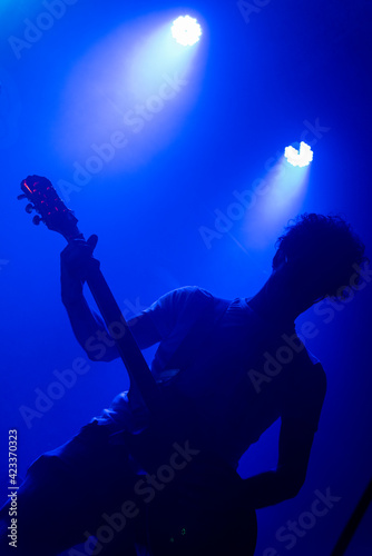 Unrecognizable male curly hair guitarrist. Music lights show silhouette during rock band live concert. Foggy light
