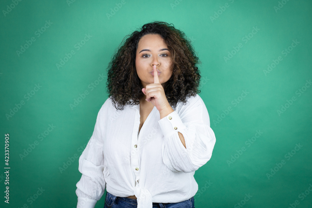 Young african american girl wearing white shirt over green background asking to be quiet with finger on lips. Silence and secret concept.