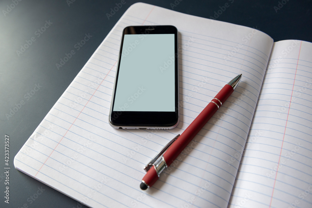 A notebook laid out flat on a blue table with a pen and a smart phone with blank screen resting on top of the ruled pages.