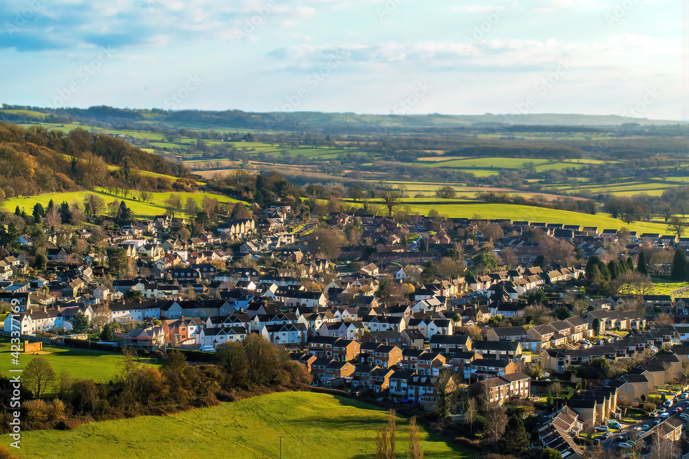 A scenic view from the top of a hill in Gloucestershire. Beautiful and colorful houses in the countryside, surrounded by green meadows and a hill.