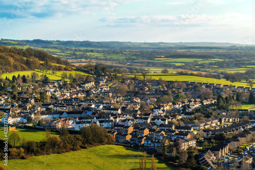 A scenic view from the top of a hill in Gloucestershire. Beautiful and colorful houses in the countryside, surrounded by green meadows and a hill.