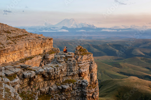 A tourist (girl) sits on the cliff edge of Bermamyt plateau against mount Elbrus and taking photos of sunset landscape. Karachay-Cherkessia, Caucasus, Russia.