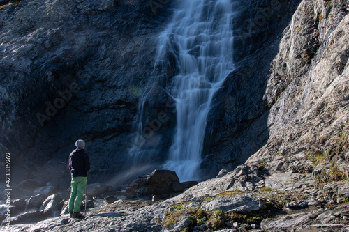 A tourist (man with trekking poles) stands in front of Sophia waterfall on sunny autumn day. Arkhyz, Caucasus, Russia.