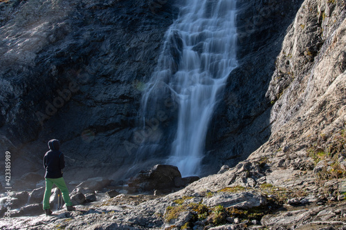 A hiker (man with trekking poles) stands in front of Sophia waterfall on sunny autumn day. Arkhyz, Caucasus, Russia.