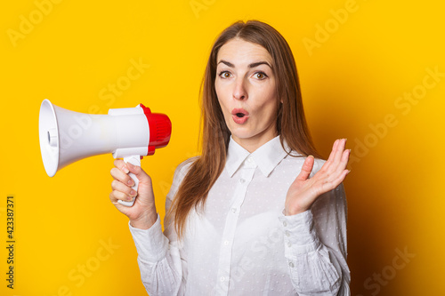 Surprised young woman in a white shirt holds a megaphone on a yellow background. Hiring concept, ad