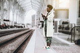 A young fancy curly-hair African woman in sunglasses, white trench, and camouflage trousers is phoning while standing on the platform of a railroad station depot and waiting for the train to travel