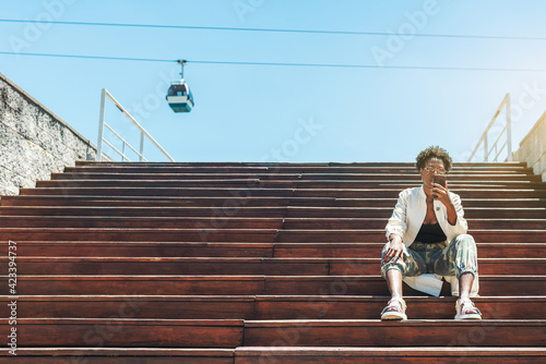 A fancy young African female is sitting on the wooden steps of the stair and using her smartphone to choose the best tourist route, a ropeway car in the background, a copy space place on the left